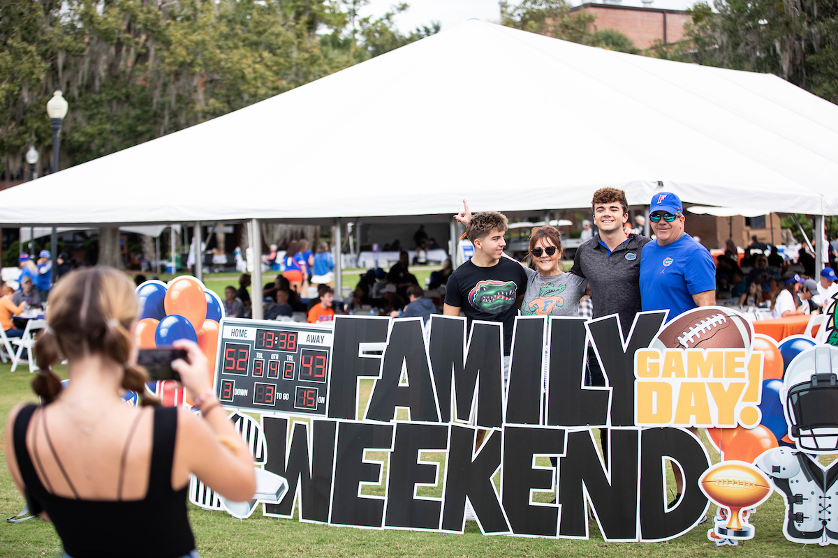 Family of four Gators fans pose behind a large, decorative yard sign that reads Family Weekend